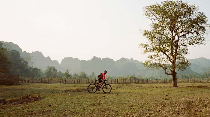 Rebecca cycling across a field in Laos. From the ride along the Ho Chin Minh Trail in 2015.
